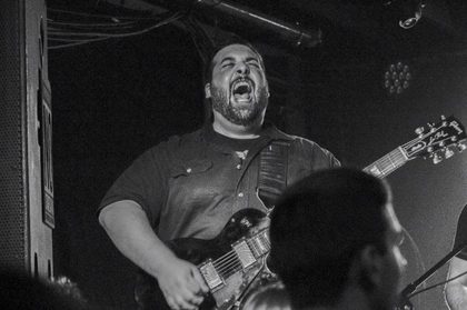 Playing guitar on a dark club stage, mid-shout, in black and white.